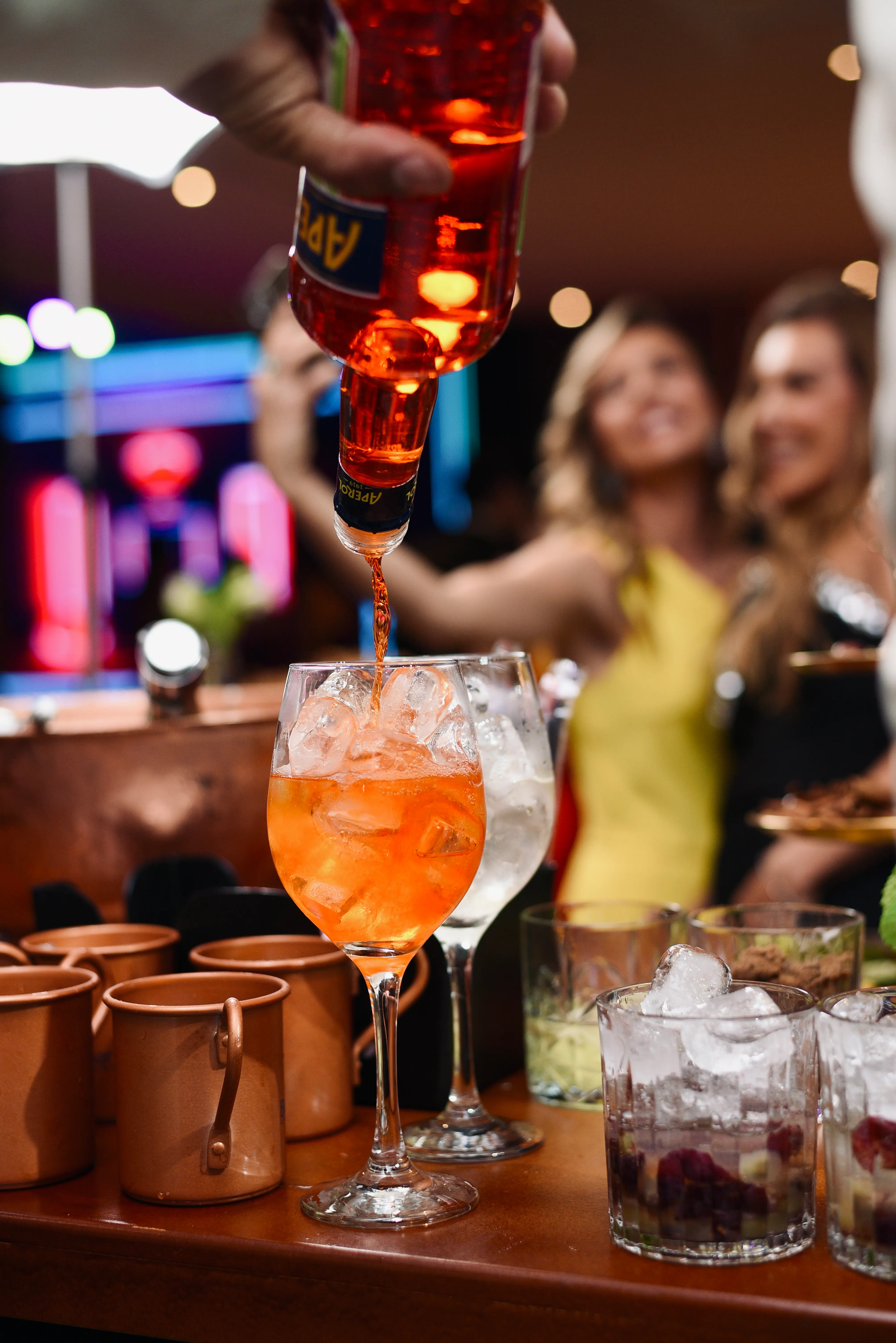 Bartender do Bar in Bar servindo um Aperol Spritz, close no copo com gelo e taças ao fundo